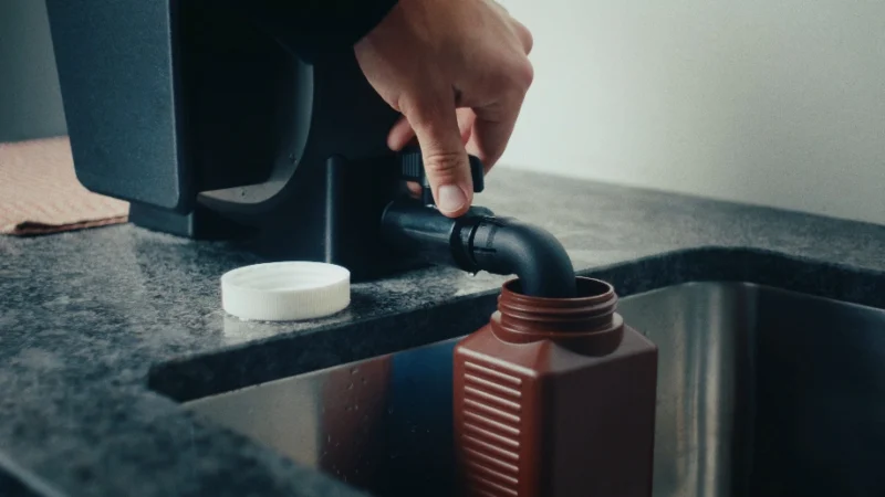 A hand turns a black spigot on a countertop appliance, dispensing liquid into a brown plastic bottle held over a sink.