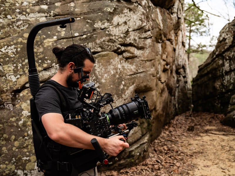 A person operates a professional video camera with stabilizing gear while standing outdoors between large rock formations on a dirt path.