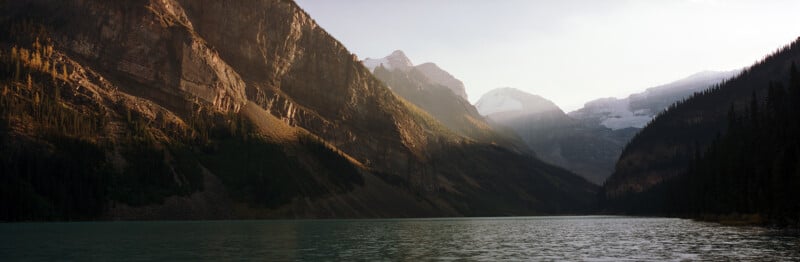 A wide mountain lake with steep rocky cliffs, pine trees, and snow-capped peaks in the background under a hazy sky.