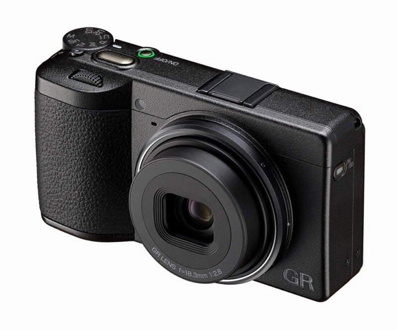A black compact digital camera with a textured grip, protruding lens, and labeled controls, positioned at an angle on a white background.