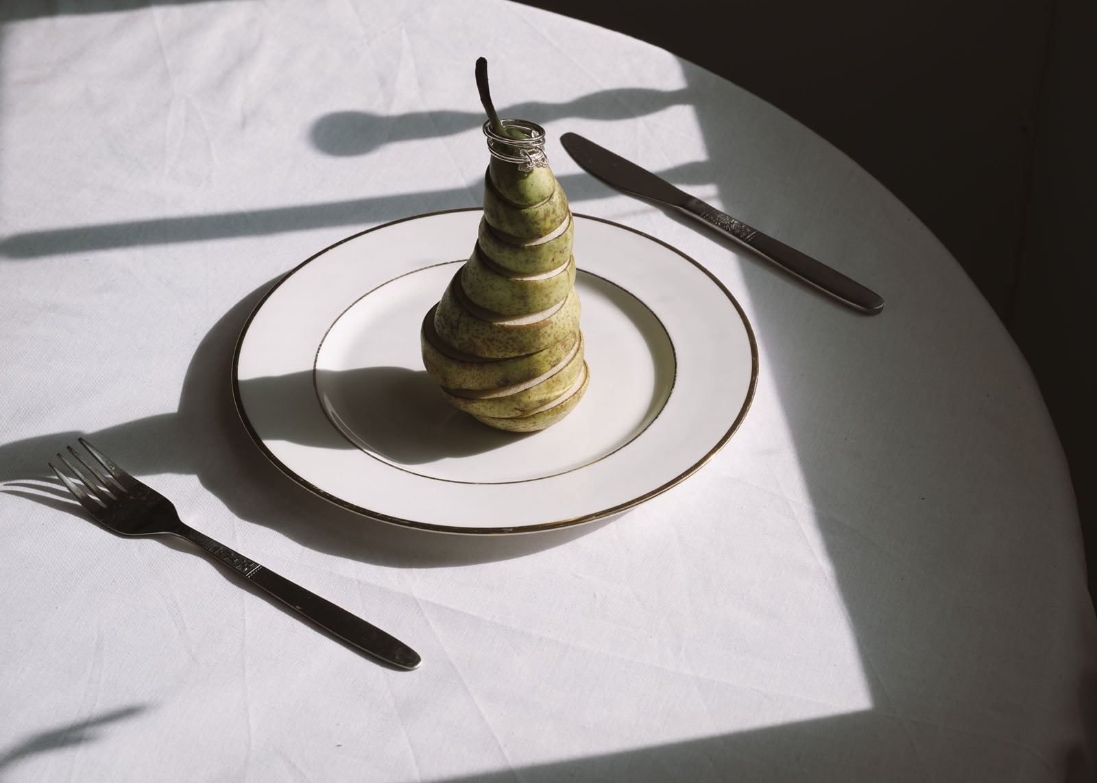A sliced pear is stacked to resemble its original shape on a white plate, set on a table with a fork and knife, with dramatic shadows cast across the scene.