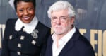 Two people pose and smile at an event. The woman wears a black dress with a white collar and floral brooch, while the man has white hair, glasses, a beard, and is dressed in a dark jacket over a white shirt.