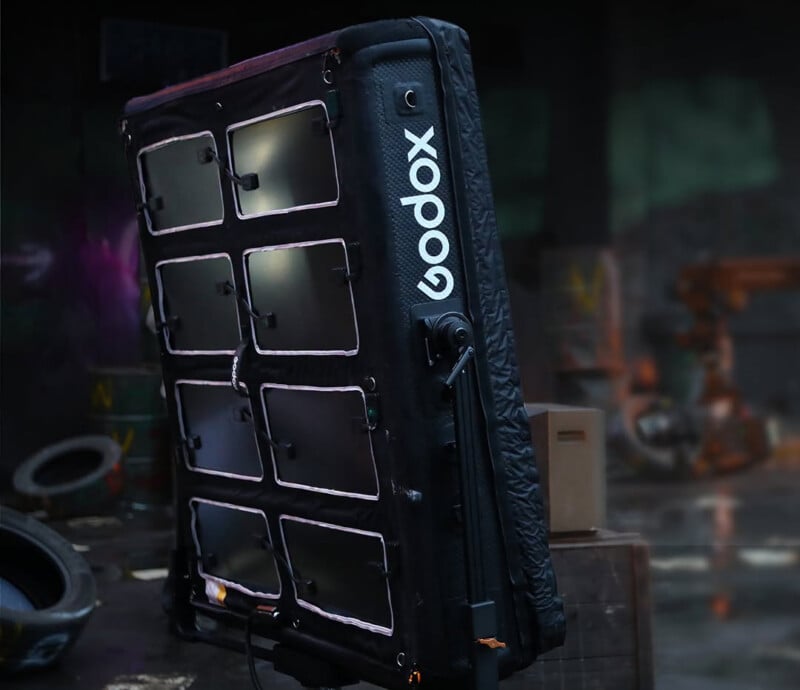 Large rectangular Godox LED panel light stands vertically in a dim, industrial setting with tires, boxes, and graffiti in the background. The panel has nine square light sections visible.