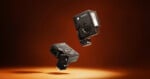 Two black compact cameras with flash attachments are suspended against a brown gradient background, with a spotlight below, giving a floating appearance.