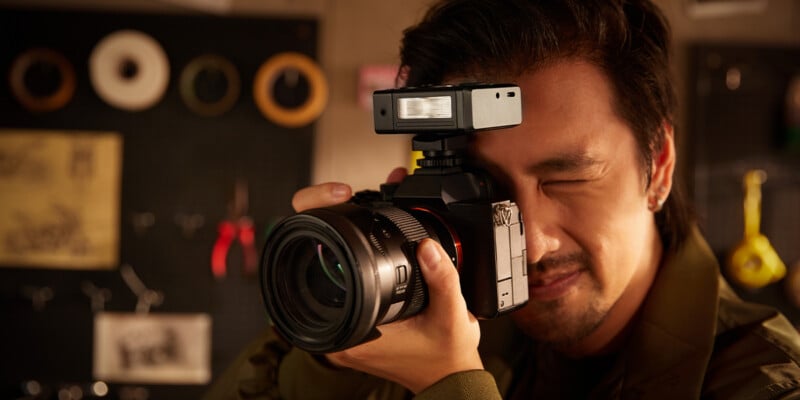 A person with dark hair is holding a professional camera up to their face, looking through the viewfinder and preparing to take a photo in a warmly lit indoor setting.