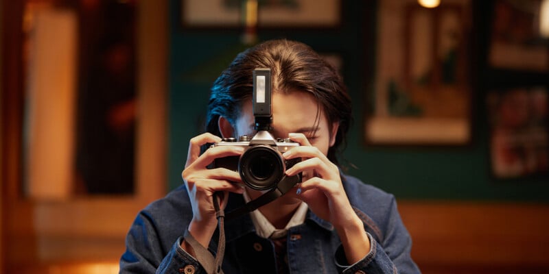 A person in a denim jacket holds a camera up to their face, preparing to take a photo in a warmly lit indoor setting with blurred framed pictures on the wall behind them.