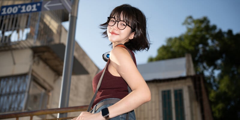 A young woman with short dark hair and glasses smiles outdoors, wearing a sleeveless top, headphones around her neck, and a smartwatch, with buildings and trees in the background.