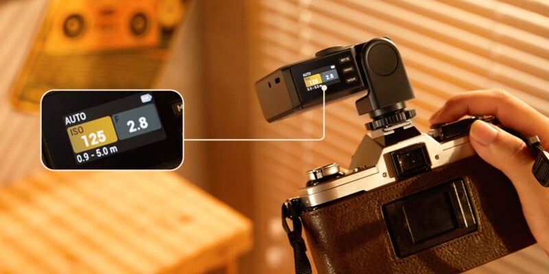 A hand holds a vintage camera with a digital light meter attached. The meter's screen displays settings: AUTO, ISO 125, 0.9-5m, and f/2.8. A close-up inset highlights the light meter's display.