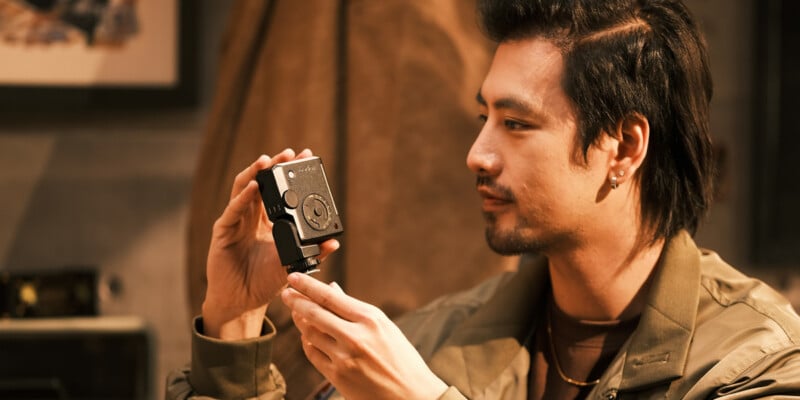 A person with dark hair and facial hair examines a small camera device indoors, holding it carefully with both hands, wearing a green jacket and an earring, with a blurred background.