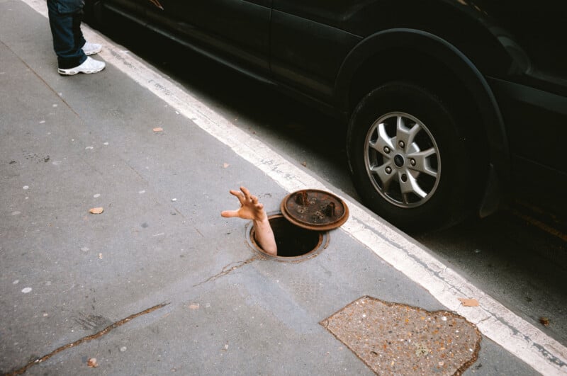 A hand reaches out of an open manhole cover in a city street next to a parked black vehicle, while a person stands nearby.