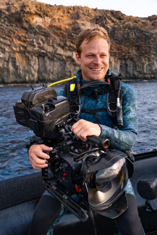 A smiling diver in a blue wetsuit sits on a boat holding a large underwater camera, with rocky cliffs and the ocean in the background.