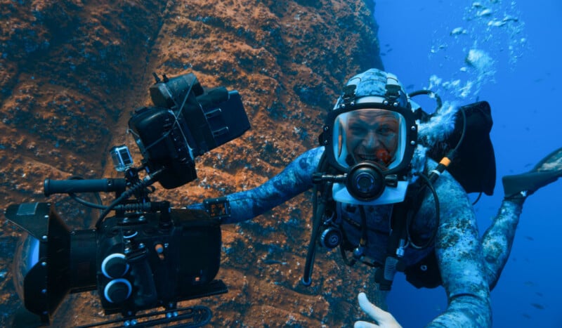 A scuba diver in a full-face mask and blue patterned wetsuit films underwater near a rocky reef, holding a large video camera as small fish swim nearby.