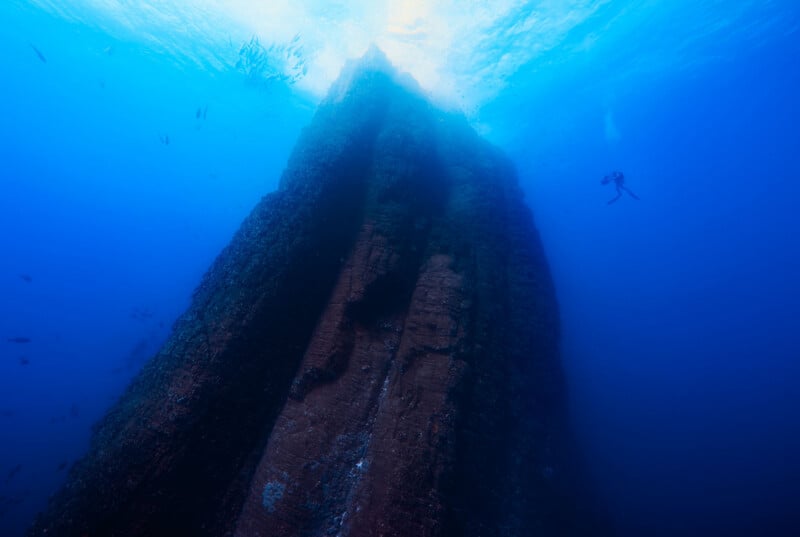 A diver swims near a towering underwater rock formation, surrounded by deep blue water and small fish, with sunlight filtering through the surface above.