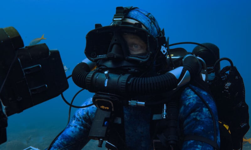 A scuba diver in a blue camouflage wetsuit and full-face mask operates underwater equipment, surrounded by blue water and a few fish in the background.