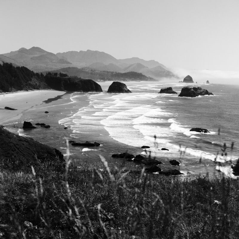 A black-and-white landscape view of a sandy beach with large rock formations in the ocean, framed by grassy hills in the foreground.