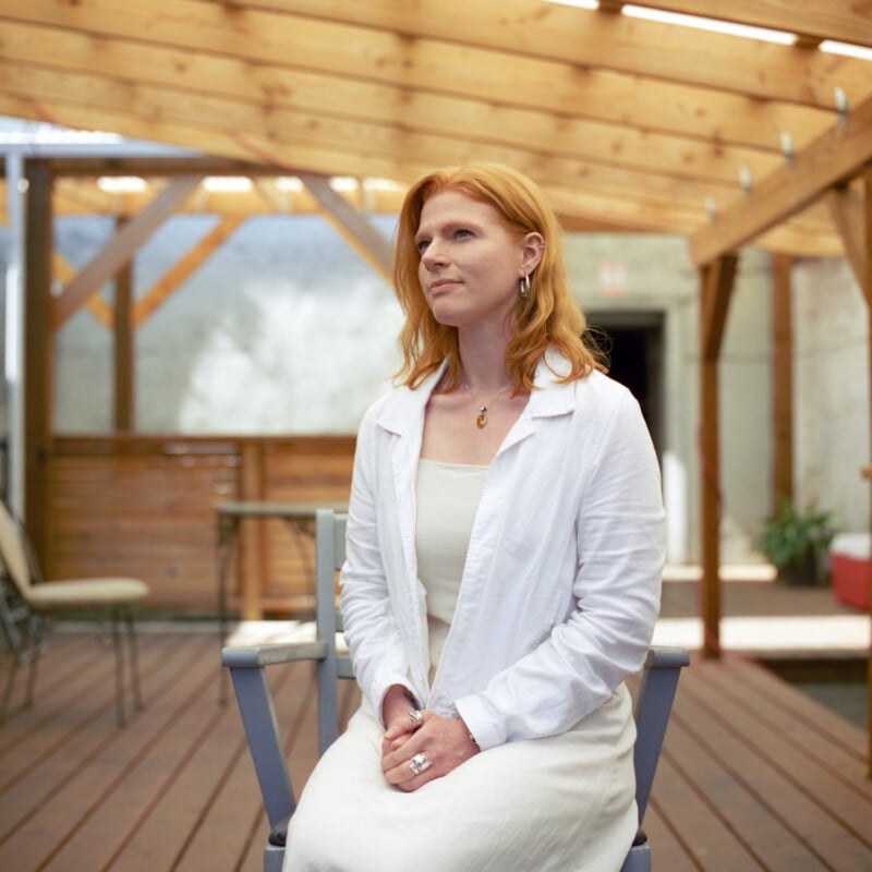 A woman with red hair, wearing a white dress and white shirt, sits on a chair on a wooden deck under a wooden roof, looking ahead with a calm expression.