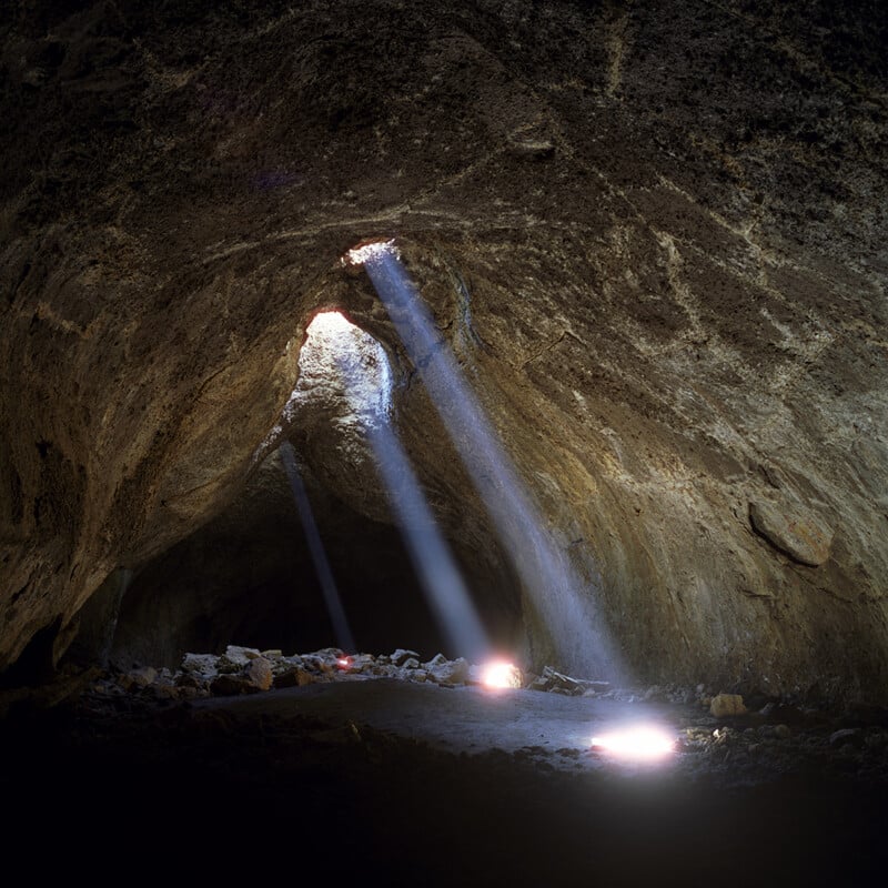 Sunlight beams through holes in the roof of a rocky cave, illuminating the cave floor with soft, bright light patches surrounded by scattered rocks and rugged cave walls.