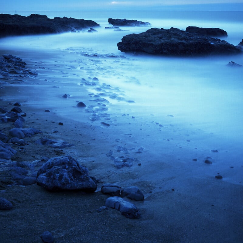 Rocky shoreline with smooth sand and misty blue water, large rocks scattered along the coast, and a calm, hazy horizon under soft light.
