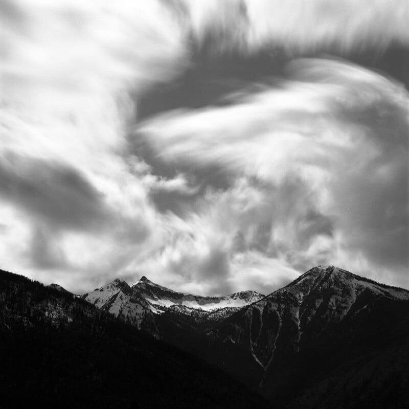 Snow-capped mountains beneath a dramatic sky filled with swirling, fast-moving clouds. The scene is in black and white, creating a moody and striking landscape.