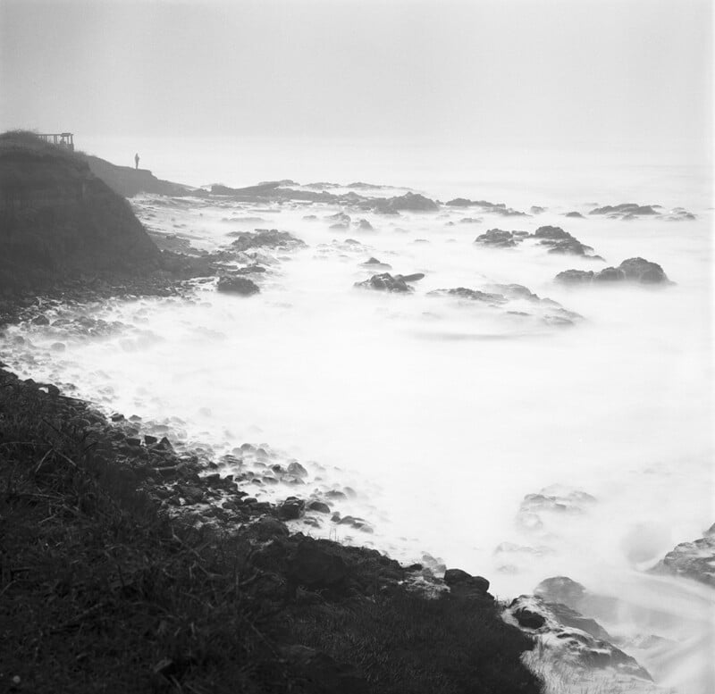 A foggy coastline with rocky shore and waves crashing. A single person stands near the edge of a cliff, silhouetted against the misty sky.