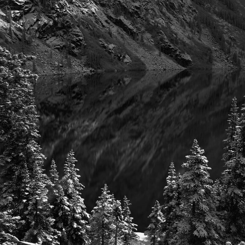 Snow-covered pine trees stand before a dark, calm lake that reflects rocky mountainsides in the background. The scene is in black and white, creating a serene and dramatic winter landscape.