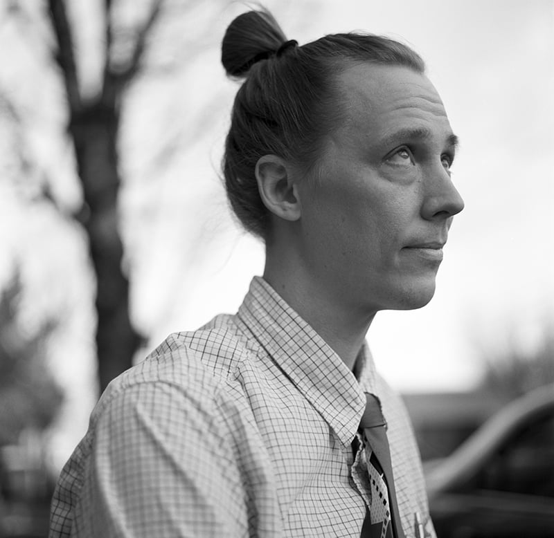 Black-and-white photo of a person with light skin, long hair in a top knot, wearing a plaid shirt and tie, looking upward thoughtfully. Blurred trees and cars are visible in the background.