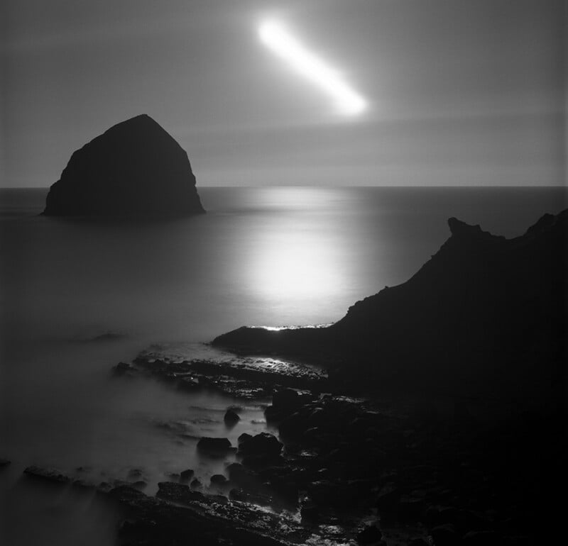 A rocky coastline at night with a large rock formation in the sea. The sky features a glowing, streaking light, reflecting on the calm water and creating a surreal, ethereal atmosphere.