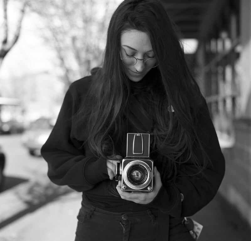 A woman with long hair and glasses looks down at a vintage film camera she is holding, standing outdoors on a city street.