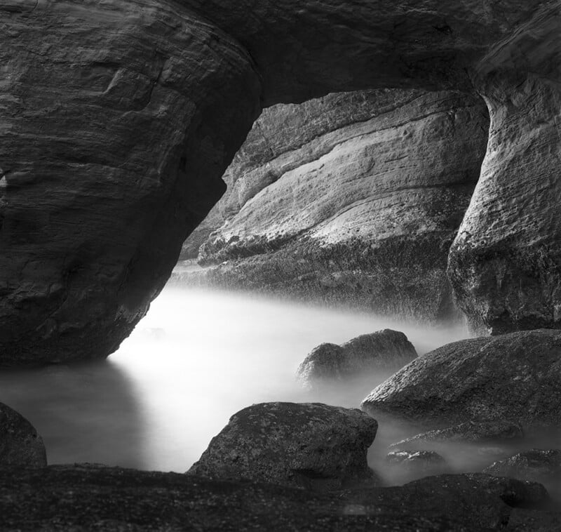Black and white photo of a rocky sea cave with smooth water flowing among large boulders. Light enters the cave from an opening, illuminating rock textures and creating a tranquil, misty atmosphere.