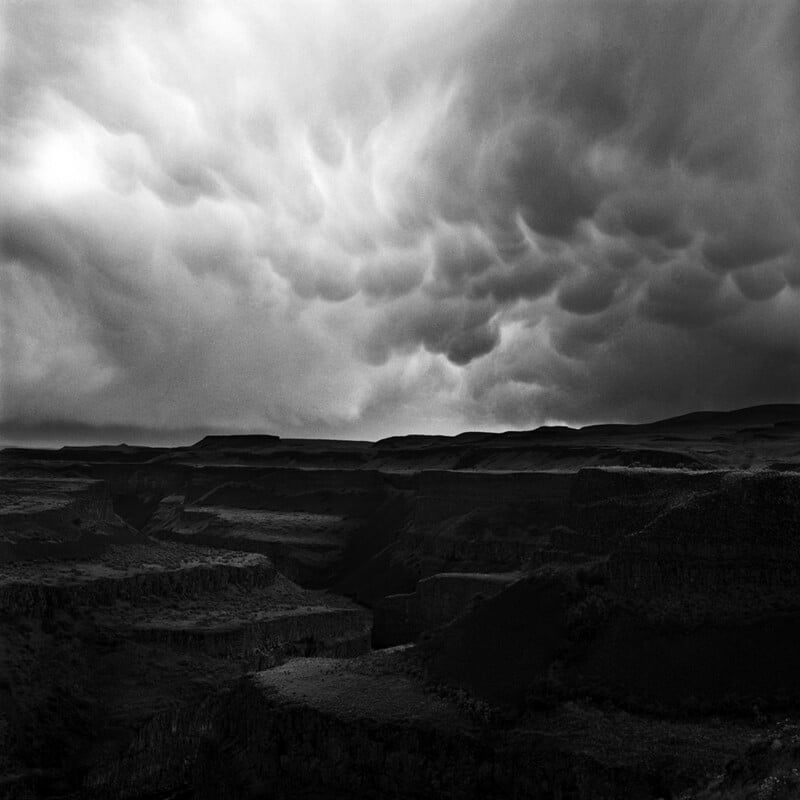 Dark, dramatic clouds loom over a rugged canyon landscape, casting deep shadows and creating a moody, atmospheric scene in black and white.