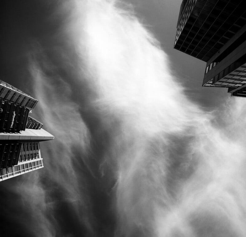 A low-angle, black-and-white view of two modern skyscrapers framing a bright, wispy cloud formation in the sky.