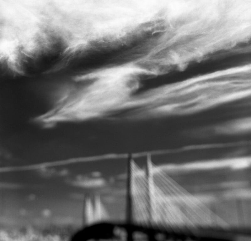 A blurred, black-and-white view of a cable-stayed bridge silhouette beneath dramatic, wispy clouds.