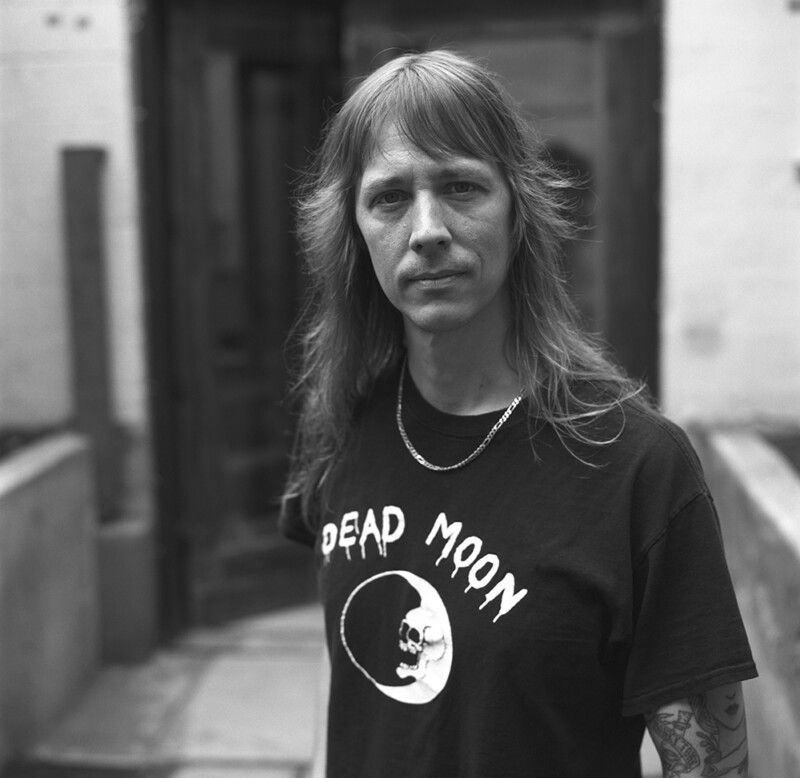 A person with long hair stands outdoors, wearing a "Dead Moon" t-shirt and a chain necklace, looking directly at the camera. The background shows a building entrance and concrete walkway. The photo is in black and white.