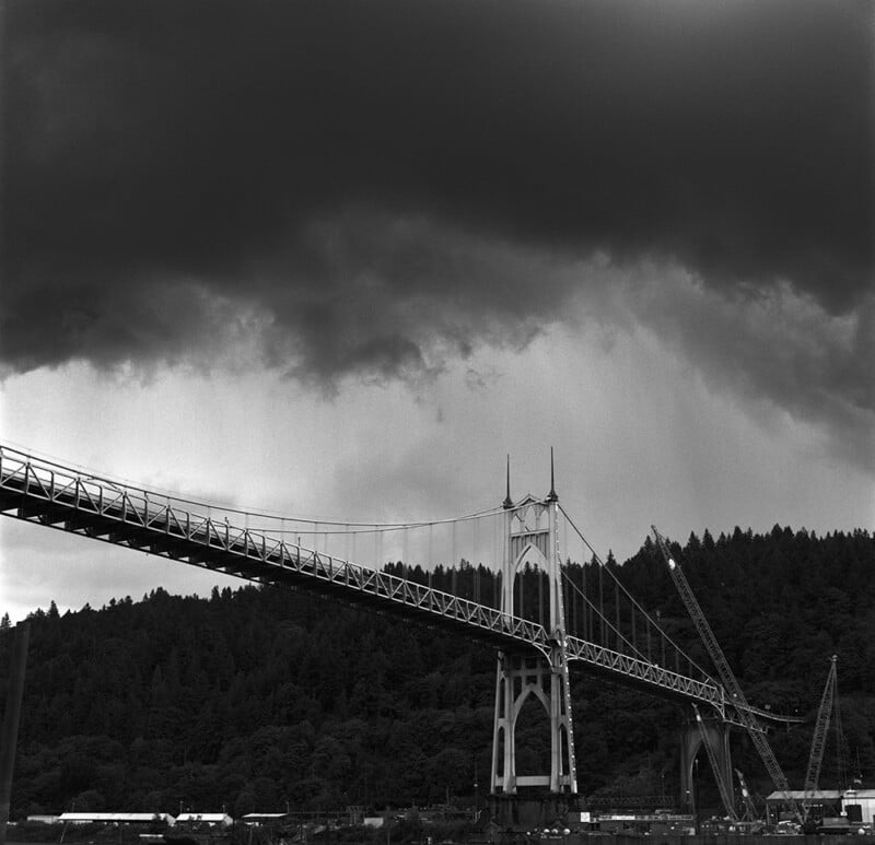 A dramatic, dark sky looms over a suspension bridge with gothic-style towers, set against a forested hillside. Construction cranes and buildings are visible at the base of the bridge.