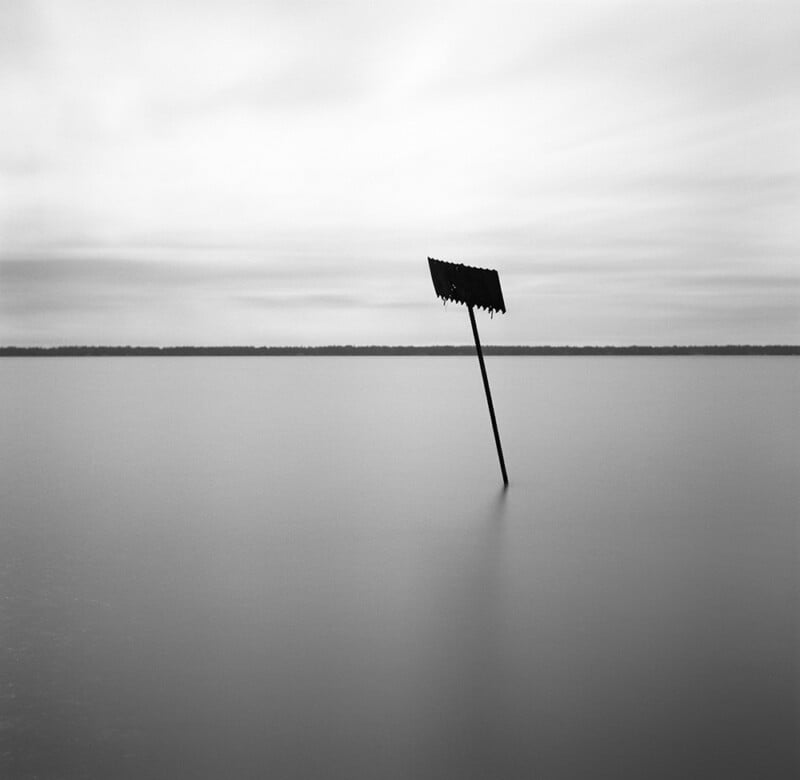 A lone, slanted signpost stands partially submerged in calm, reflective water under a cloudy sky, with a dark, distant shoreline stretching across the horizon.