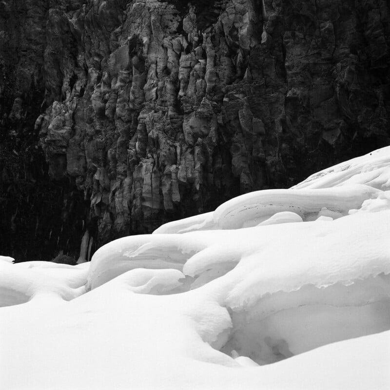 Snow-covered rocks or ice formations in the foreground with a rugged, dark rock cliff face in the background, captured in black and white. The scene appears cold and dramatic.