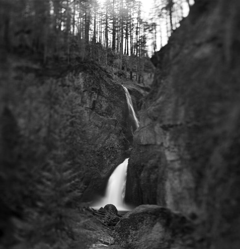 Black and white photo of a tall waterfall cascading down rocky cliffs, surrounded by trees. Sunlight filters through the forest at the top of the image, creating a serene, natural scene.