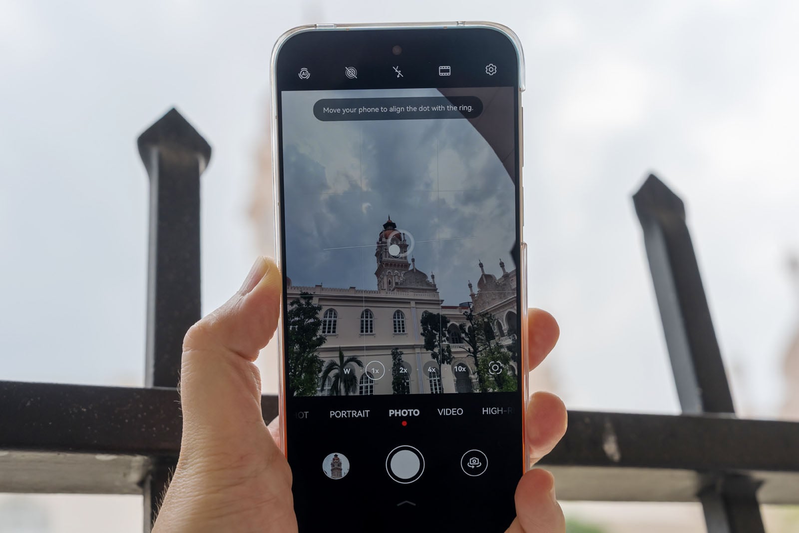 A hand holds a smartphone, using its camera to photograph a historic building with a clock tower through a black metal fence.