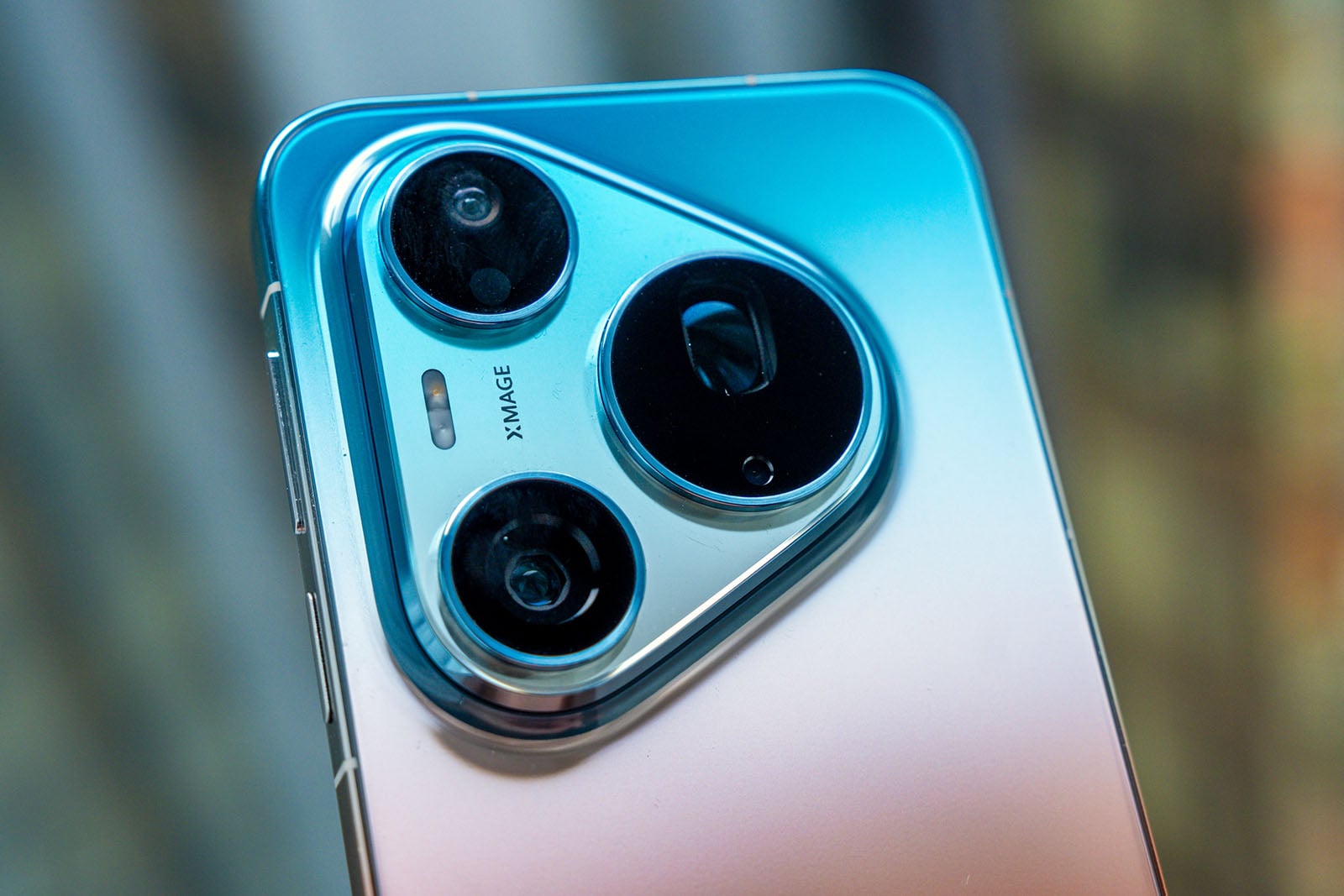 Close-up of a smartphone’s rear camera module with three lenses and a metallic blue gradient finish. The camera area is triangular with rounded corners and metallic edges. Background is blurred.