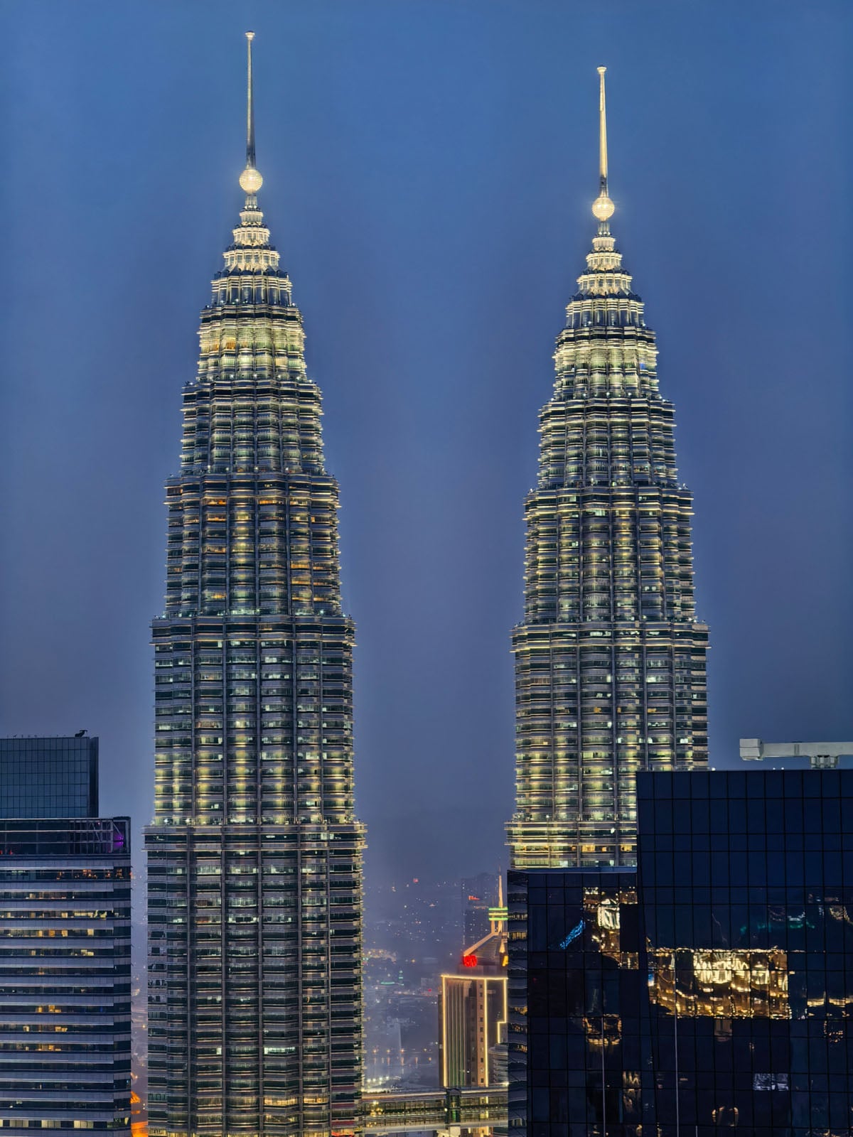 The image shows the illuminated Petronas Twin Towers at night in Kuala Lumpur, Malaysia, with surrounding buildings partially visible in the foreground.