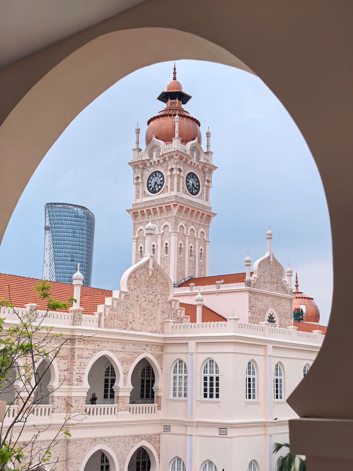 A historic clock tower with a copper dome rises above a grand white building, framed by an arch. A modern glass skyscraper stands in the background against a cloudy sky.
