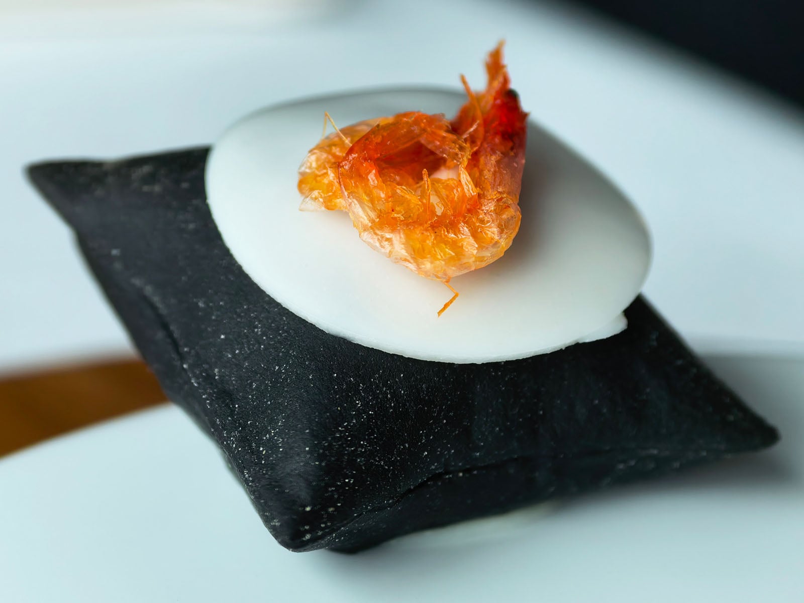 A crispy shrimp sits atop a white circular base, which is placed on a small black pillow-like cushion, creating a modern, artistic food presentation.