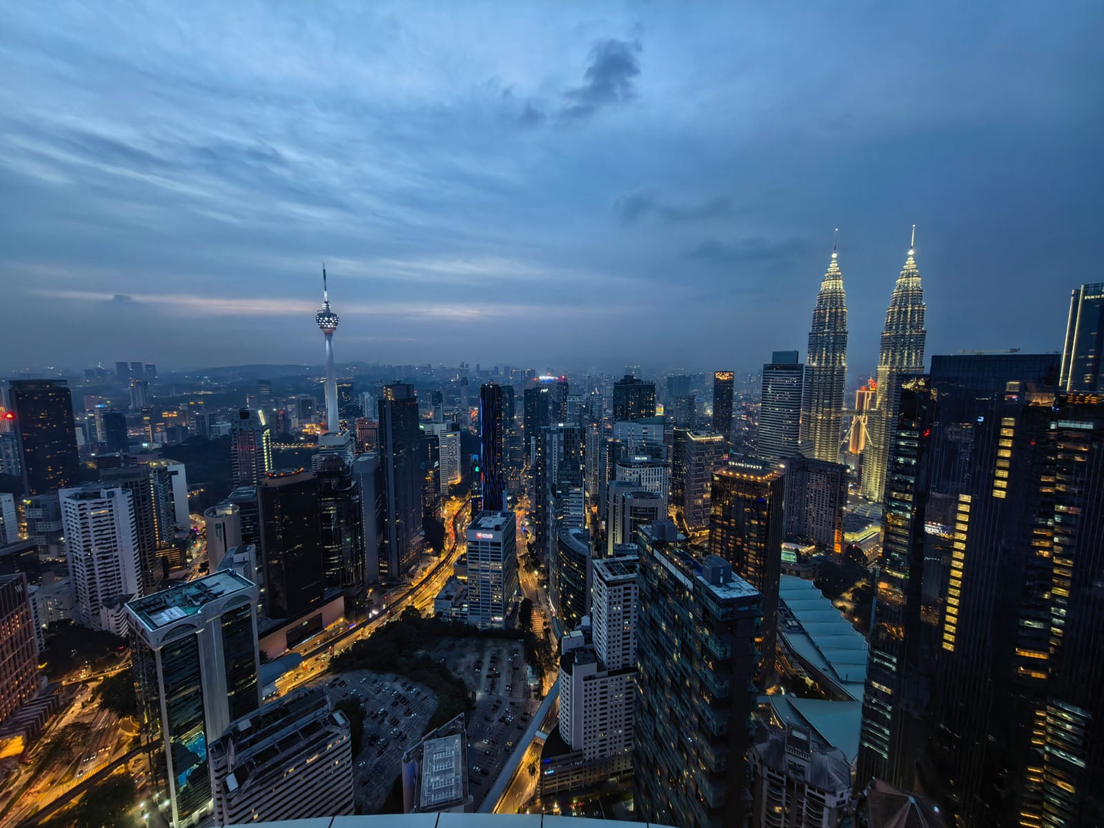 Aerial view of a city skyline at dusk with illuminated skyscrapers, including twin towers and a tall communications tower, under a blue, cloudy sky. Streets and buildings are lit up, revealing an active urban landscape.