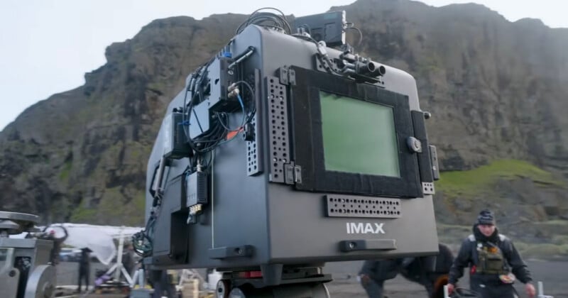 A large IMAX camera is set up outdoors with rocky hills in the background. Film crew members and equipment are visible around the scene.