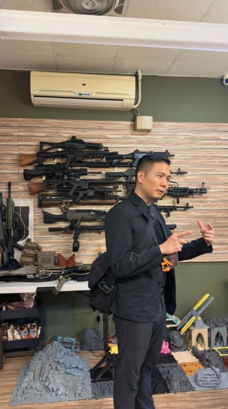 A man in a black blazer and backpack stands in front of a wall display of various rifles and tactical gear.