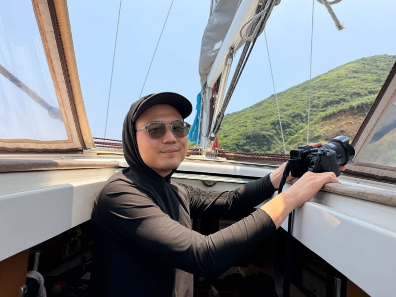 A man wearing a black hooded shirt and sunglasses holds a camera while sitting on a sailboat.