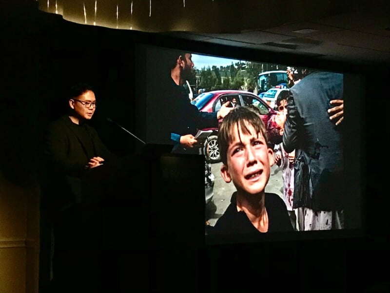 A man in glasses and a dark blazer stands at a podium, speaking in front of a large screen displaying a photograph of a crying boy amidst a scene of conflict.