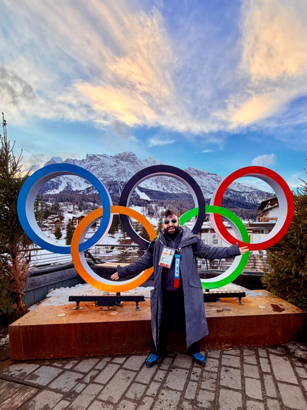 Person in winter clothes stands smiling in front of large Olympic rings statue, with snowy mountains and buildings in the background under a colorful sunset sky.