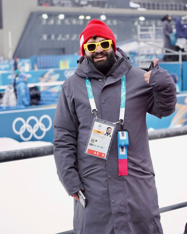 A person in a gray coat, red beanie, and yellow sunglasses stands by an Olympic venue, smiling and giving a thumbs up. They wear accreditation badges and there is snow in the background.