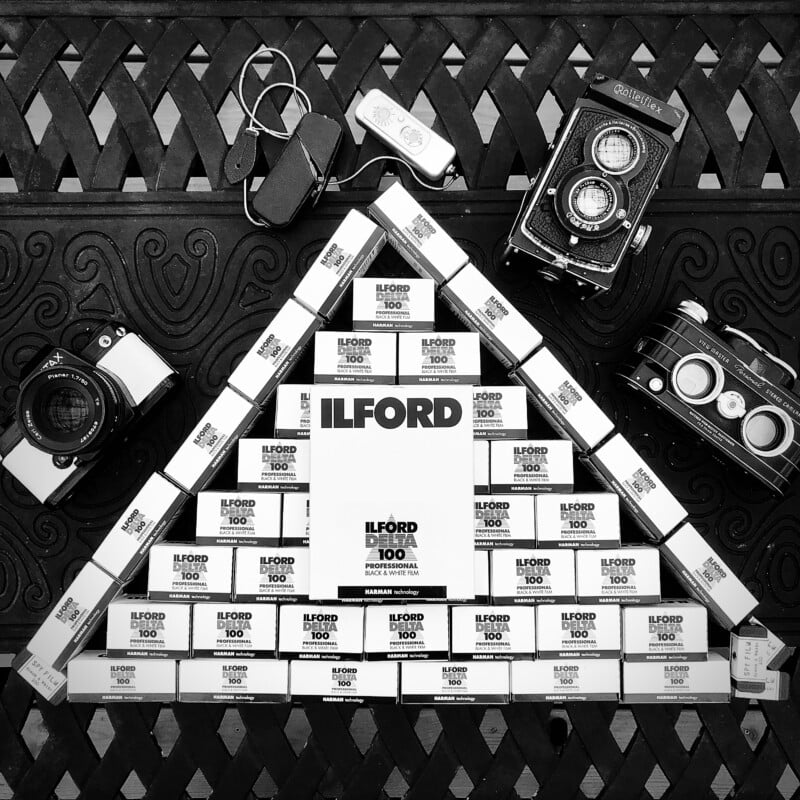 A pyramid of Ilford Delta 100 film boxes is surrounded by four vintage cameras and two light meters, all arranged on an ornate patterned surface in a black-and-white photograph.